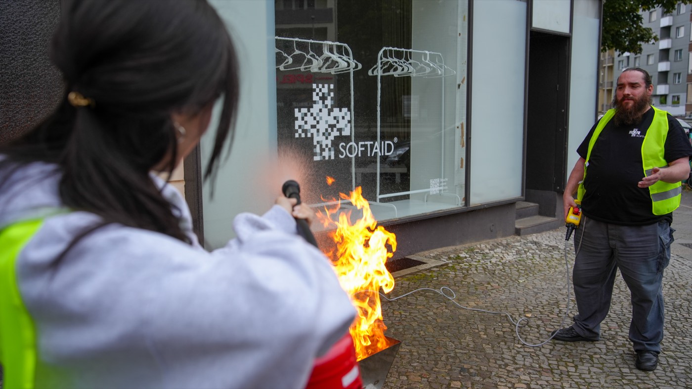 Eine Teilnehmerin in Warnweste löscht das Übungsfeuer, daneben der SoftAid-Trainer